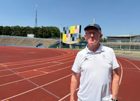 A man in a white Team GB T-shirt stands on Crystal Palace track with the iconic scoreboard in the background covered by netting.