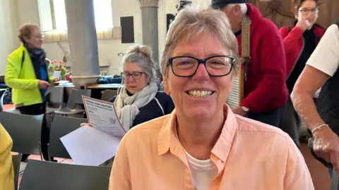 A woman sits in a church during a public meeting, smiling. She is wearing a coral coloured shirt with a white T-shirt underneath and black-rimmed glasses. She has short, blonde hair. There are people sitting behind he and walking to their seats.