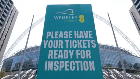 Getty Images A large turquoise sign that tells people to have their match tickets ready for inspection. It is at the bottom of a wide staircase that leads towards Wembley Stadium.
