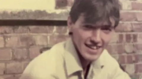 Family photograph Undated image of Jonathan White with brown hair wearing a white jacket in front of a brick building. He is smiling.