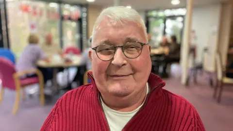 A man looking at the camera and smiling. He is wearing a white t-shirt under a red, knitted jumper.