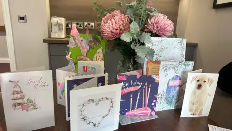 A display of birthday cards arranged on a wooden table, including one with a puppy and another featuring candles on a cake. A vase of pink flowers sits behind the cards. Two handmade cards reading 'Happy 106 Birthday' are visible at the edges of the table.