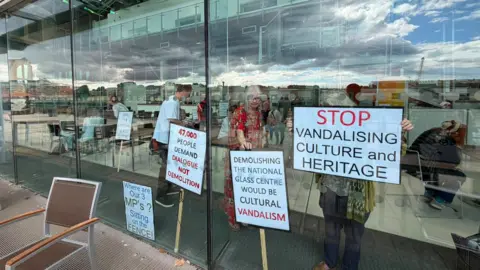 Campaigners seen through the glass front of the building with multiple placards. The first on the right reads 'Stop vandalising culture and heritage', the next 'Demolishing the National Glass Centre would be cultural vandalism', then to the left there is '47,000 people demand dialogue not demolition' and finally on the far left 'Where are our 3 MP's? Sitting on the fence.' Due to the reflection it is hard to see how many people are inside, but it looks to be at least 10. Cranes, buildings and clouds are reflected in the glass.