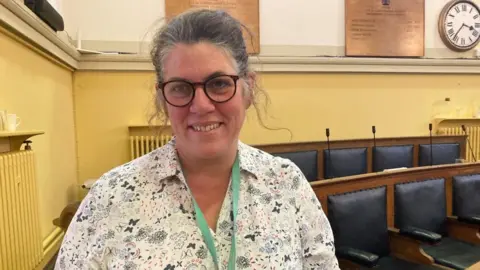 A woman is standing in a council chamber. She is wearing floral top and glasses and is smiling at the camera. There are empty seats to her left and a clock in the far right corner.