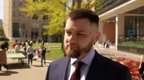 A man with a beard and short brown hair is wearing a dark-coloured suit with a white shirt and red tie. He is standing outdoors with a group of people in the background who appear to be socialising near a lawned area.
