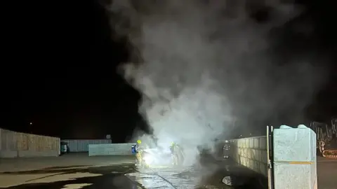 Smoke bellowing from a vehicle fire outdoors with two firefighters stood either side with hoses. There is a large area with walls on the left and right. The vehicle is central and the ground around it is wet. 