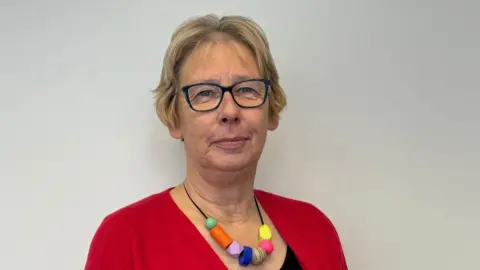 A woman wearing a red cardigan with a black top. She is also wearing glasses and a beaded necklace. She is standing against a light-coloured backdrop and is smiling for the photograph.