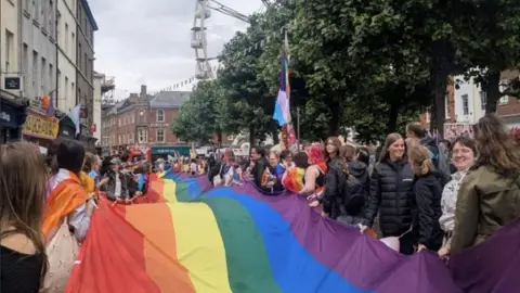 Crowds of people in York holding a giant rainbow flag, with some holding flags and wearing coats