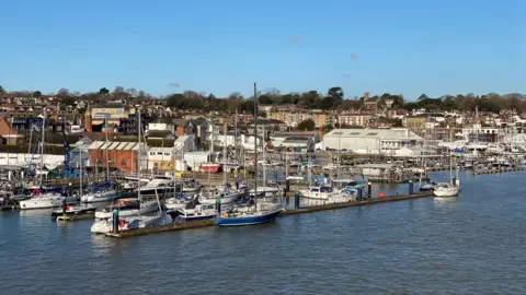 Caz of Wight A view over a town from the sea. In the foreground are several moored yachts. Behind you can see industrial buildings and homes. The sky overhead is blue. The photo appears to have been taken from a boat. 