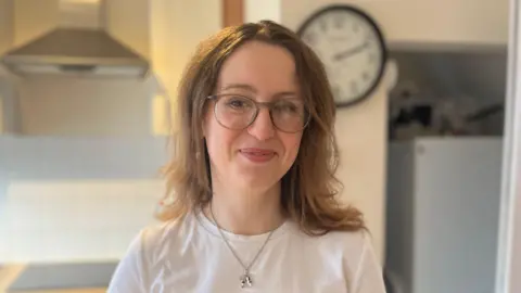 Susie stands in a kitchen wearing a white T-shirt. The kitchen has light-coloured cabinets, a stainless steel oven with a stovetop, and a metallic range hood. A black-and-white clock is mounted on the wall near a refrigerator in the background.