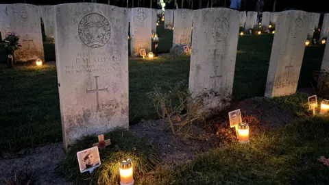 Venray War Cemetery Adoption Graves Foundation Close up image of graves at the British war cemetery in Venray, Netherlands. All the graves have a small bright lantern on the ground in front of them and most have a photograph of the soldier buried there. 