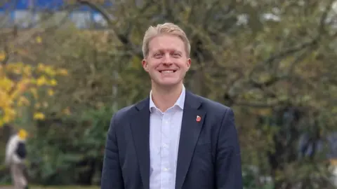 Dr Ben Spencer smiles at the camera, he is wearing a dark blue suit jacket with a white shirt underneath with the top button undone. In the background out of focus trees are seen