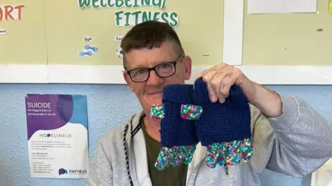 A man in his 60s wearing glasses and a grey sweatshirt is holding up a pair of navy and multicoloured spotty gloves. The background shows a display reading 'wellbeing and fitness' and has a suicide prevention helpline poster on the left. 