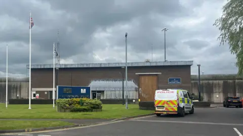 Nondescript prison block building with a police van outside. It is a grey day. There are several white poles in front of the building. 