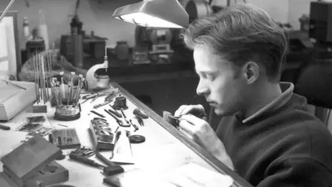 Roger Smith A black and white image of a young Roger Smith working at a work bench. He's on the right and on the desk are various fine tools under a desk lamp. He's using hand tools to do intricate work on a watch.