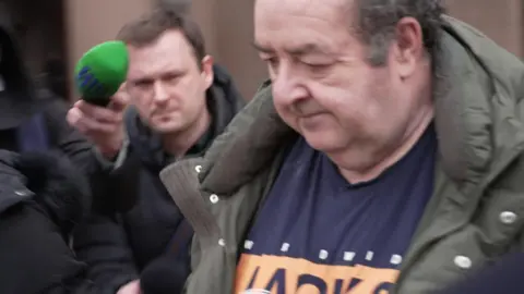Gerard Woodhouse, who is wearing an olive green overcoat and a Jack & Jones t-shirt, is seen leaving court. He has his head down and is surrounded by reporters, some with microphones. 