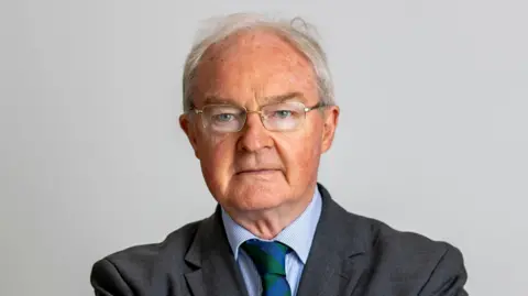 A man with white hair looking at the camera, he has glasses and dark blazer, blue shirt and green and blue tie