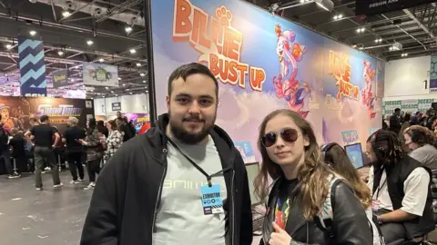 A man in a hoody and a woman wearing sunglasses stand in a convention centre hall