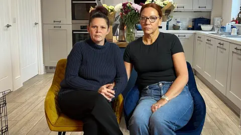 Renee Turner, left, has dark hair tied back and is wearing a navy jumper, black trousers, and is sitting on a yellow chair. Ruth O'Loughlin, right, has brown hair tied back, glasses, a black shirt and jeans and is sitting on a dark blue chair. They are both in a kitchen with units and cupboards behind them.