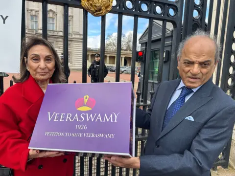 A woman in a red coat holding a big purple cardboard box outside the gates of Buckingham Palace with 'Veeraswamy 1926, petition to save Veeraswamy" wrriten on it. Also holding the box is a man in a suit. 