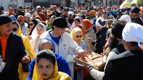 Gurmit Sandhu and Ranjit Sahota A parade of hundreds people in traditional Sikh head coverings in a street. A man in a black turban with long black beard holds out a box of pizza in the foreground, which a young man takes a slice from.