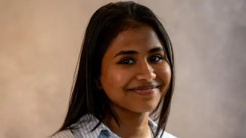 Sithuni Mallawaarachchi is smiling at the camera and is wearing a blue and white striped shirt.
