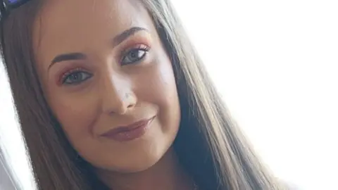 Chloe Mitchell is smiling in the picture. She has part of her hair tied up in a high bun while the rest is straightened. She is wearing makeup. She also has sunglasses on her head.