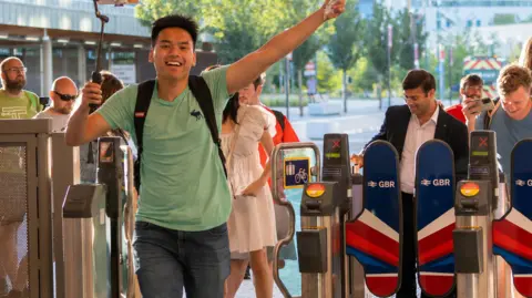A man wearing a green shirt and dark jeans. He is holding a selfie stick in his right hand and his left hand is in the air. He appears to be running with a big grin on his face. Behind him are barriers and several people standing behind them