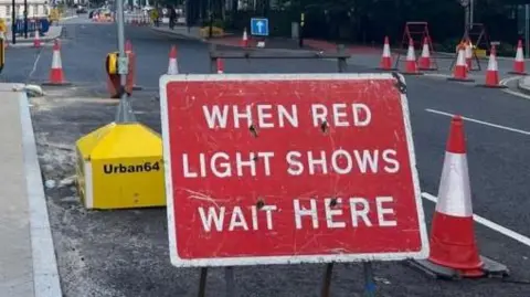 A sign reading "when red light shows wait here" placed on a central lane of Cumberland Place, Southampton with a temporary traffic light showing green. Bollards line the road to close off the lane to traffic.