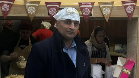 Sanjeev Kumar, wearing a black jacket, blue shirt and white cloth around his head. He is giving a slight smile to the camera. Behind him are people wearing aprons dishing up food. He has red and gold bunting above his head.