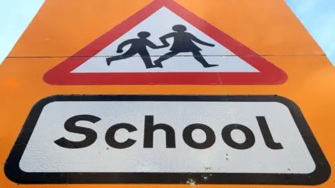 A picture of a yellow sign. It says the word school on it with a graphic above it.