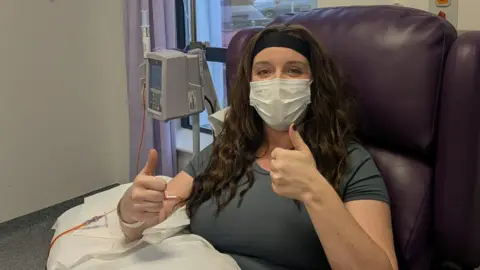 Other A woman in a chair, wearing a mask, gives a thumbs up sign to the camera. She wears a grey top, has long brown hair past her shoulders and a black hairband. Over her lap is a white sheet and she sits in a dark purple, padded chair. 