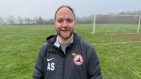 Adam Slowman wearing a Nike branded Fairford Youth FC windbreaker. a grass pitched goal area with a muddy surface can be seen in the background.