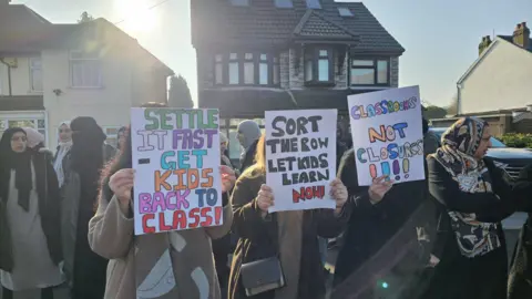 Parent Voice A group of men, women and children all wearing coats are stood at the side of the road holding placards. Many of their faces are hidden