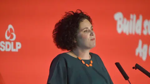 Claire Hanna. She has dark curly hair, a dark green top and orange necklace. She is standing in front of microphones. A red background is behind her with SDLP branding on it.