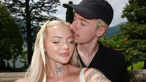 Handout A woman with long blonde hair has her eyes closed and is smiling as a man wearing a black baseball cap kisses the top of her head. There are trees behind them.
