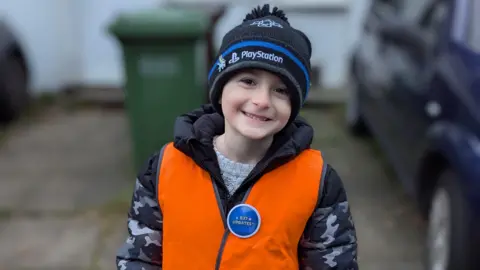 Harlee is smiling at the camera. He is wearing a black and dark blue hat, with a grey and black camo coat, and an orange hi-vis jacket.