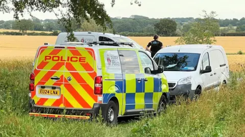 Stuart Woodward/BBC Three police vans parked on a field, with an officer looking away into the field.
