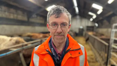 Ian Cursiter, a grey haired bespectacled man wearing a bright orange jacket smiles at the camera in his cow shed