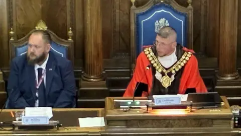A man with a partially shaved head and glasses is wearing a bright red and black robe, with a white frill and large gold chain. He is sitting next to a man with brown hair and a brown beard in a navy blue suit with white shirt and navy tie. They are sitting at a large wooden desk in a council chamber