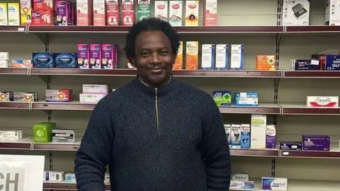 Tunde Olawoye stood behind the counter at his pharmacy wearing a blue fleece jumper. There are shelves of drugs and medicine behind him.