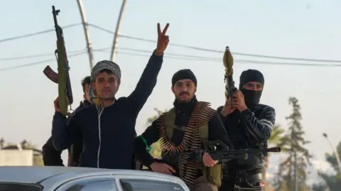 Getty Images Three young men with weapons are standing in the back of a truck. One has an RPG, the other a heavy machine gun. The man in the foreground holds an AK47 rifle and raises his fingers in salute.