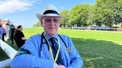 Tom Williams/BBC Colin Roberts standing outside next to a course at Newmarket racecourse. He is looking directly at the camera and smiling. He is wearing a blue shirt with a blue tie and cream hat. Round his neck he has a yellow lanyard and stethoscope on.