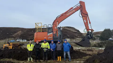 Six men standing in front of a large red digger. They are surrounded by mounds of earth in a rough looking landscape. Two of the men are wearing high-vis jackets and four of them are wearing blue hats. There are vehicles and a small hill in the background.