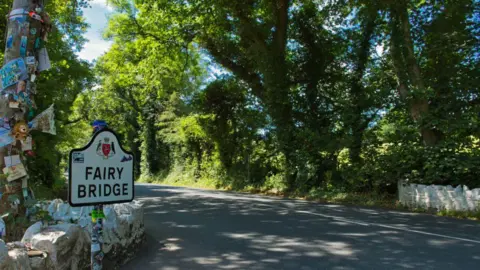 MANX SCENES A sign reads fairy bridge you can see the white bridge on either side of the road you can see large arching trees on either side of the road.