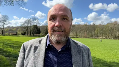BBC Head and shoulders of Dr David Scott. He has a short beard flecked with grey ad is wearing a dark blue shirt and grey jacket. He is standing at the site of the massacre. It's a beautiful day with the sun shining and a lush green field with trees at the edges behind him.