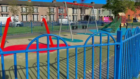 Gill Burdekin A children's playground in a park. Blue painted railings are in the foreground. A large red frame with two chains on the end is in the centre frame, it is empty, with no swing attached. A traditional swing-set is next to this.