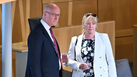 Getty Images A bald man with glasses wearing a black suit, white shirt and purple tie is standing beside a woman with blonde hair who his wearing a white jacket and black and white blouse. She has sunglasses on her head and both are holding glasses of water.