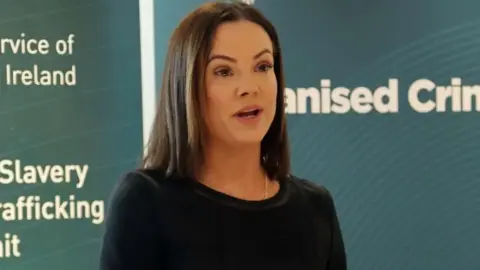 Pacemaker Det Insp Rachel Miskelly has long brown hair and is wearing a black top. She is standing in front of PSNI banners.