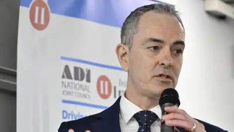ADINJC Stewart Lochrie, chairman at the Approved Driving Instructors National Joint Council. He has short salt and pepper hair and is wearing a navy blue suit and tie and white shirt. He is speaking into a hand-held microphone.